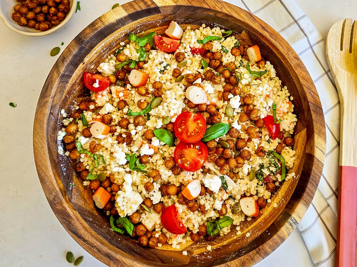 Vue de dessus d'un grand saladier en bois rempli d'une salade de perles de blé composée de couscous perlé, pois chiches rôtis, feta émiettée, tomates cerises coupées, dés de surimi et basilic frais, posé sur une nappe blanche à rayures avec des couverts en bois sur le côté.