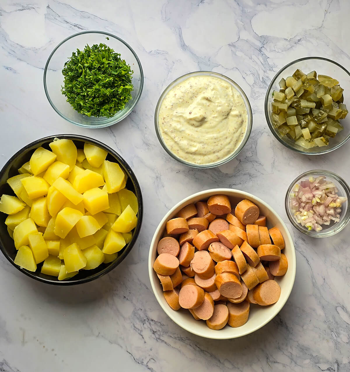 ue de dessus d'un plan de travail en marbre blanc avec six bols contenant les ingrédients de la salade strasbourgeoise : pommes de terre bouillies en cubes, rondelles de saucisses de Strasbourg, dés de cornichons, échalotes émincées, persil haché et un bol de sauce à la moutarde à l'ancienne.