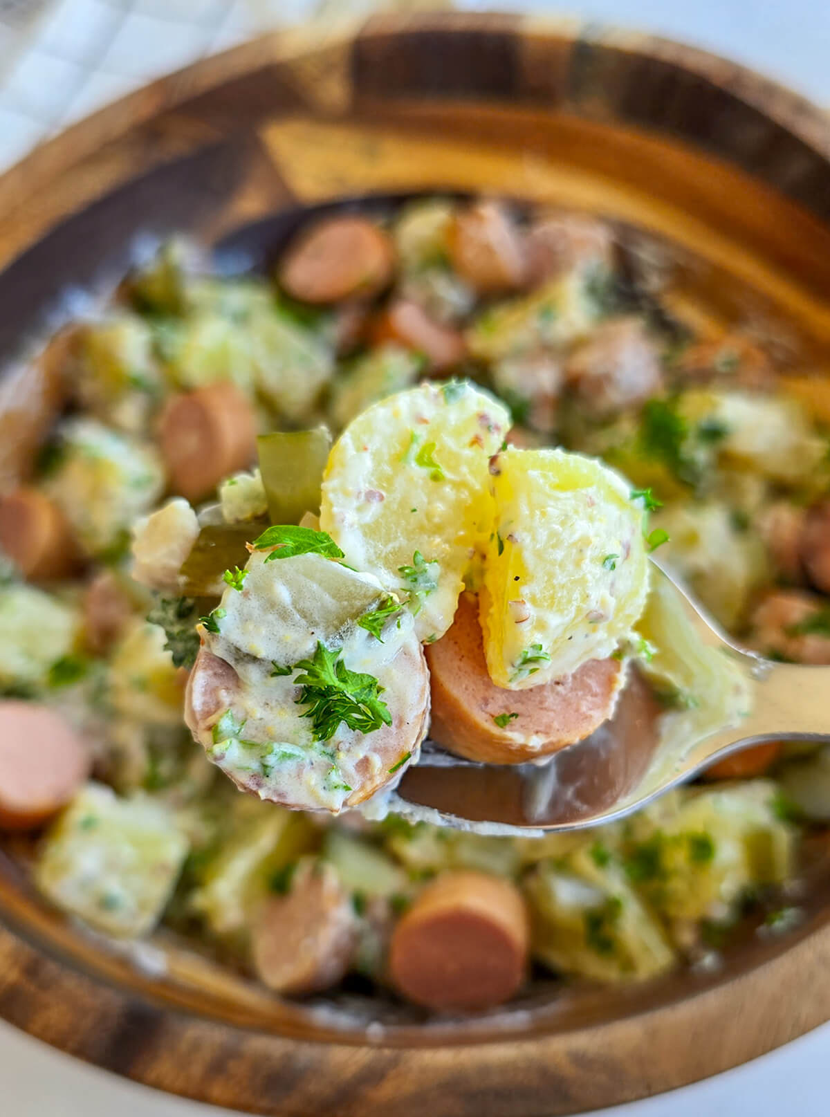 Gros plan de dessous d'une cuillère en acier inoxydable contenant une bouchée de salade strasbourgeoise, avec des pommes de terre crémeuses, des morceaux de saucisse et des cornichons, maintenue au-dessus du bol principal en bois flou.
