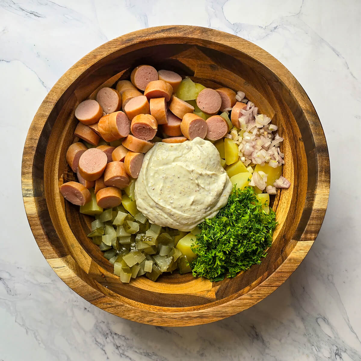 Plan moyen de dessus d'un bol en bois contenant des tas distincts d'ingrédients pour la salade strasbourgeoise (saucisses, pommes de terre, échalotes, persil, cornichons), au centre desquels se trouve une généreuse louche de sauce crémeuse