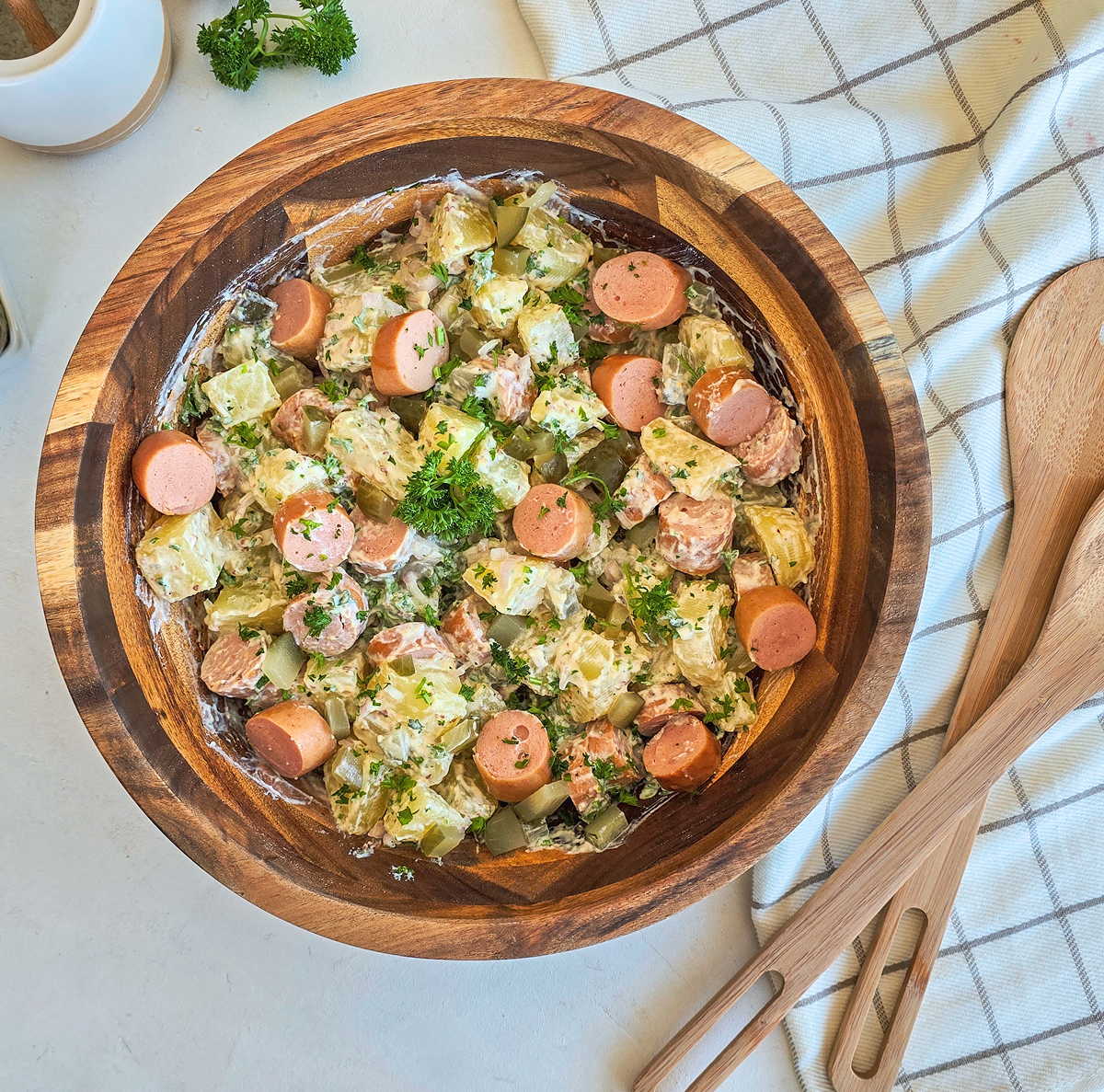 Vue de dessus d'un grand bol en bois rempli d'une salade strasbourgeoise. sur une table blanche, à côté d'un torchon quadrillé gris et blanc et de couverts de service en bois.