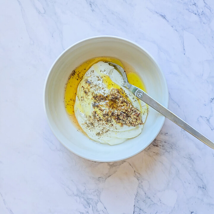 Préparation d'une sauce salade crémeuse dans un bol blanc contenant du skyr ou yaourt, de la moutarde à l'ancienne, de l'huile d'olive et du poivre.