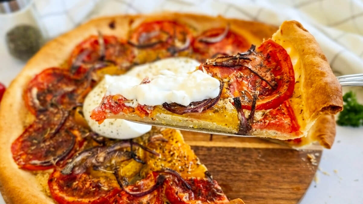 Gros plan sur une part de tarte à la tomate, moutarde et oignons rouges soulevée à la spatule, garnie d'une cuillérée de stracciatella ou burrata crémeuse.