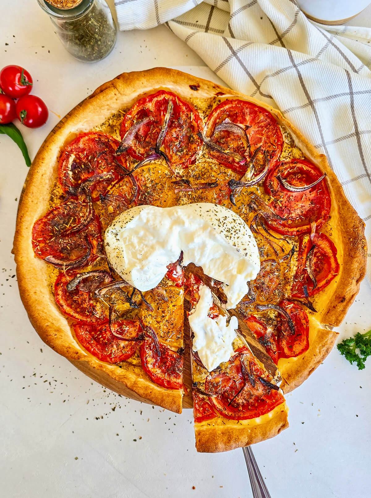 Une part de tarte fine aux tomates et oignons rouges, surmontée de burrata crémeuse, est soulevée au-dessus du plat posé sur une table en marbre blanc.