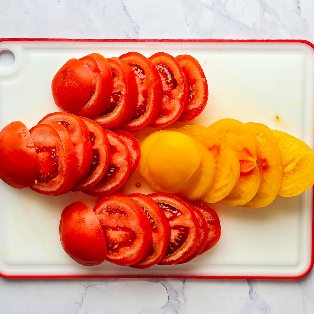 Des tomates coupées en rondelles sur une planche à découper blanche, sur un fond blanc marbré.