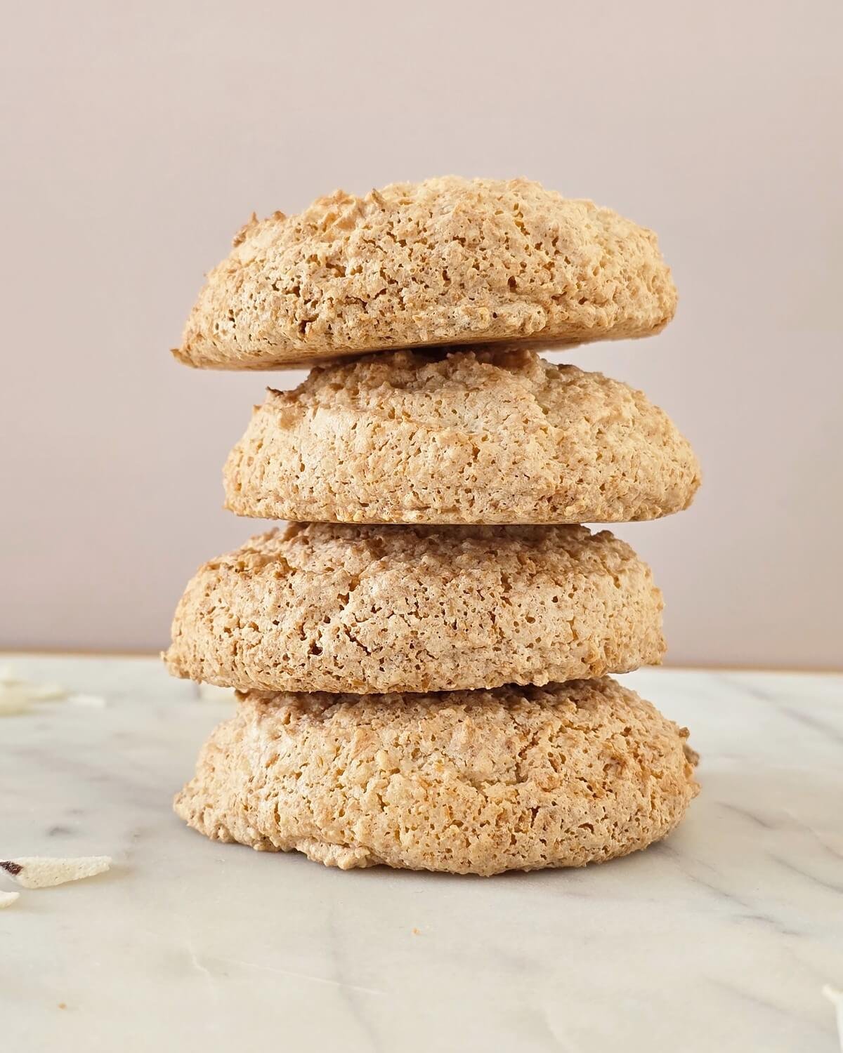 Pile de quatre biscuits à la noix de coco dorés et croustillants, posée sur un plan de travail en marbre blanc devant un fond rose pâle.