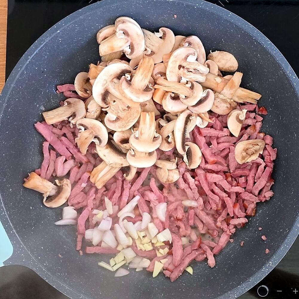 Lardons, champignons de Paris émincés, échalote et ail qui commencent à revenir dans une poêle pour la garniture des patates douces farcies.