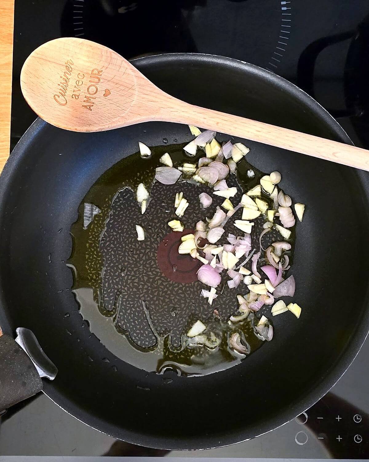 Échalote et ail émincés qui revenent dans l'huile d'olive au fond d'une poêle noire, avec une cuillère en bois.