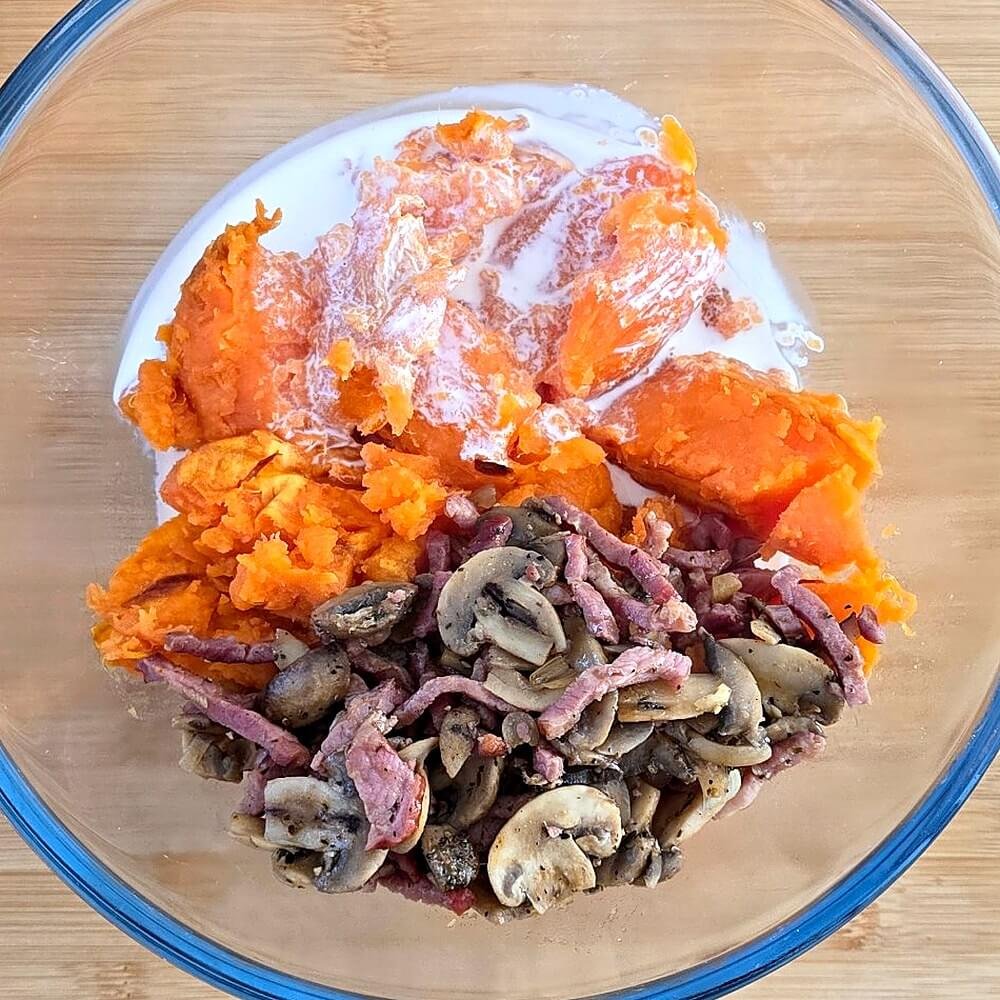 Saladier en verre avec la chair écrasée des patates douces, la crème et la garniture de lardons et champignons, prêts à être mélangés pour la farce.