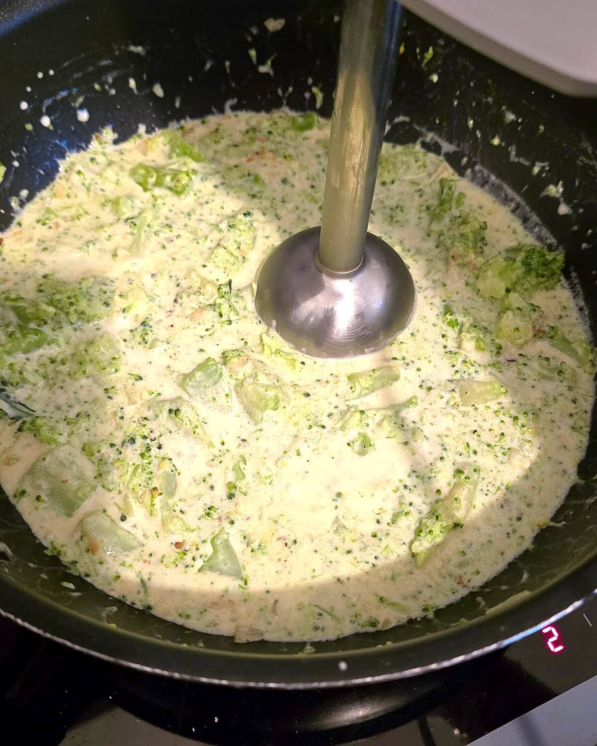 Mixeur plongeant dans une poêle de crème et de brocoli, en train de transformer le mélange en sauce onctueuse pour les pâtes.