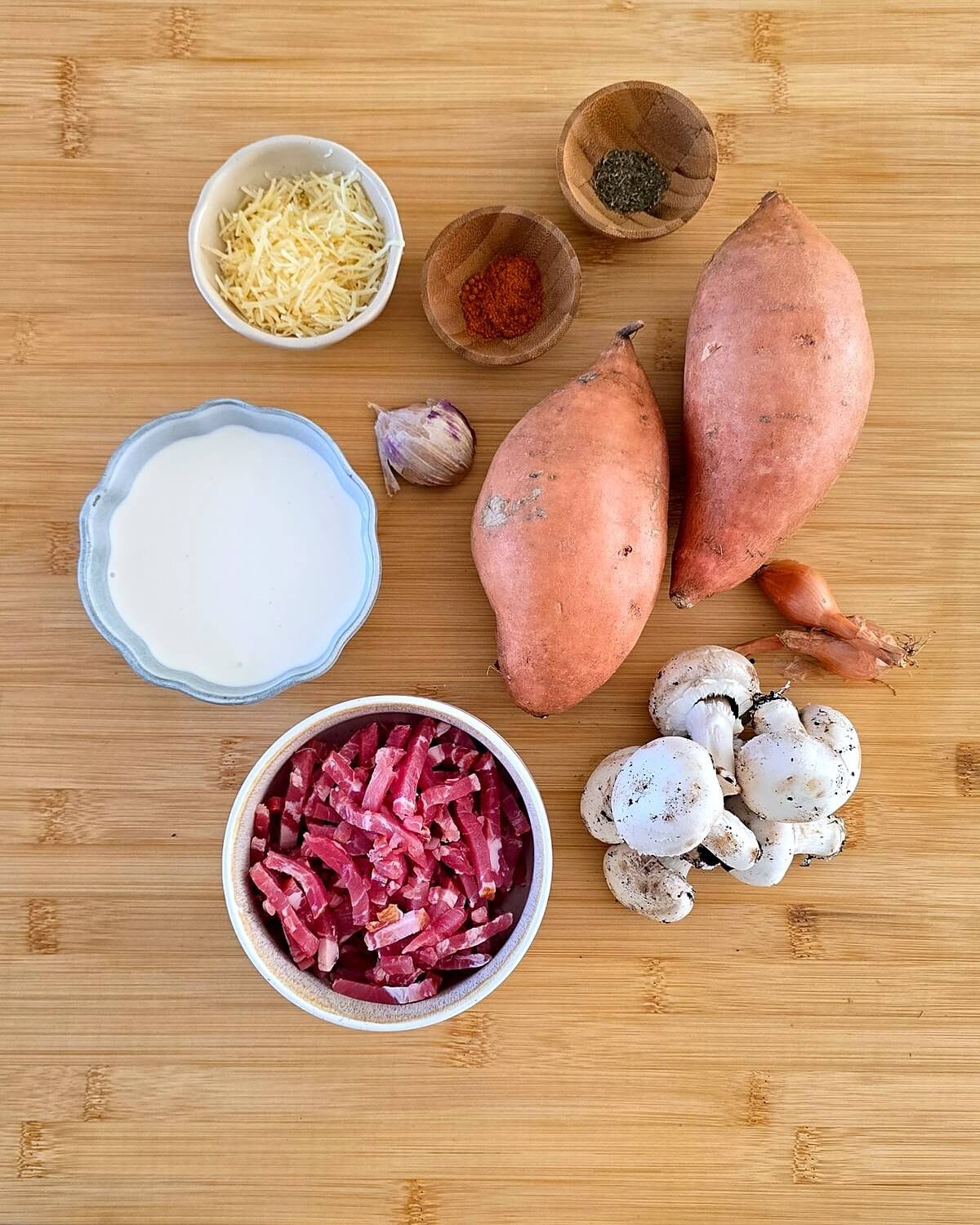 Ingrédients d’une recette à la patate douce disposés sur une planche en bambou : deux patates douces, des lardons, de la crème, du fromage râpé, du paprika, des herbes séchées, une gousse d’ail, des échalotes et des champignons de Paris.