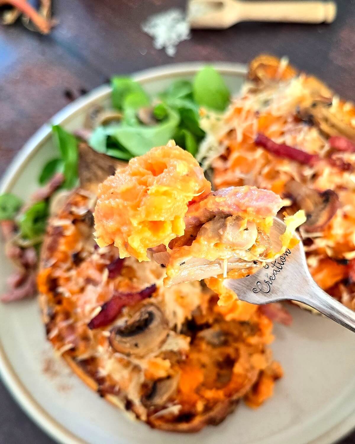 Deux patates douces farcies aux lardons et champignons, gratinées au fromage râpé, servies avec de la mâche dans une assiette beige sur une table en bois foncé.