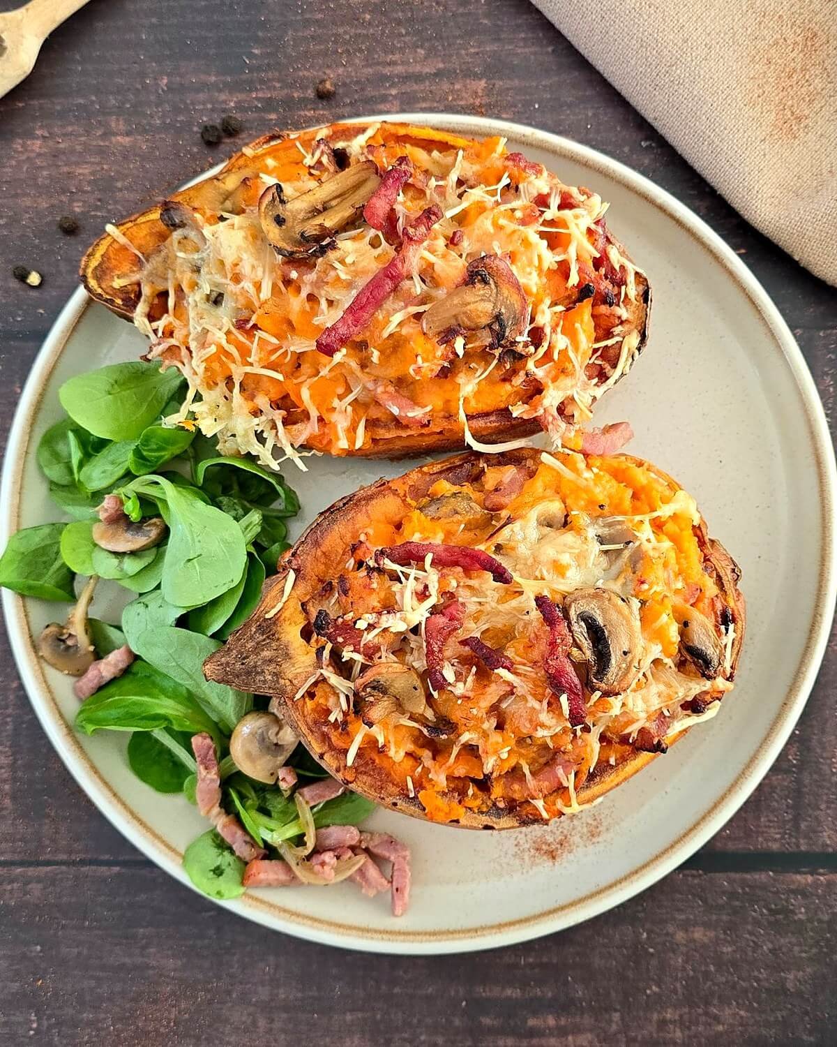 Deux patates douces farcies aux lardons et champignons, gratinées au fromage râpé, servies avec de la mâche dans une assiette beige sur une table en bois foncé.