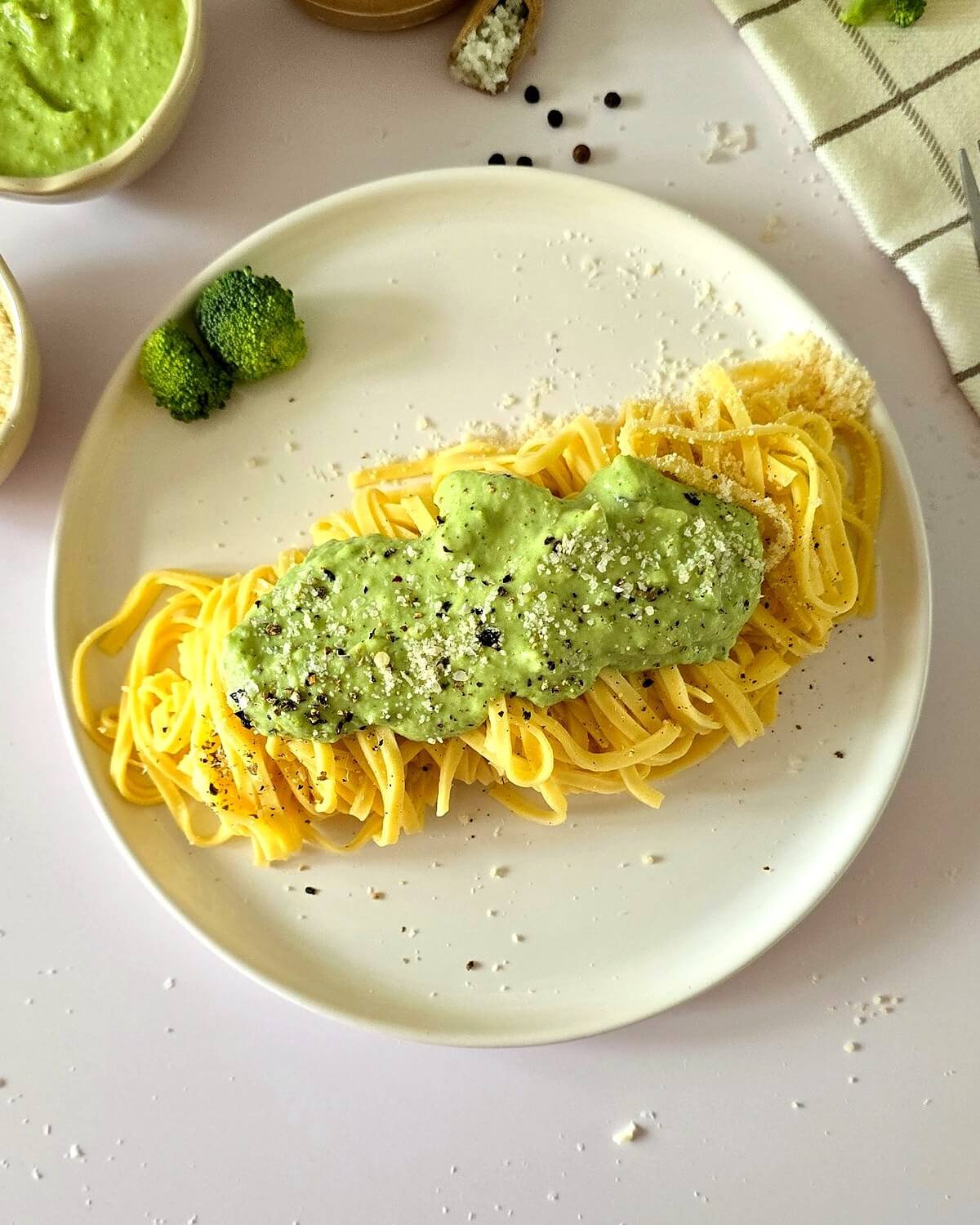 Pâtes au brocoli à la crème sur une assiette blanche, avec du parmesan et de la crème de brocoli sur les bords, le tout sur un fond blanc avec un torchon à carreaux.