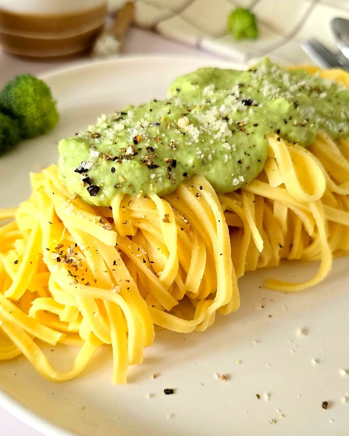 Gros plan de tagliatelles nappées d’une sauce crémeuse au brocoli, saupoudrée de parmesan râpé et de poivre noir concassé, servies dans une assiette blanche.