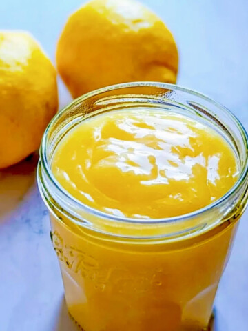 Pot de lemon curd onctueux fait maison avec deux citrons frais en arrière-plan.