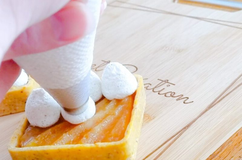 Une main pochant de la crème chantilly vanille sur une tartelette tatin sur une planche de bois