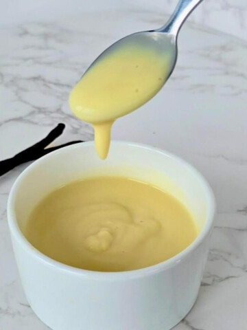 Crème anglaise onctueuse à la vanille coulant d'une cuillère en argent au-dessus d'un ramequin blanc.