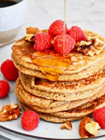 Des pancakes avoine banane dans une assiette beige, avec un filet de sirop d'érable et des framboises fraiches.