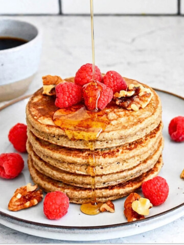 Des pancakes avoine banane dans une assiette beige, avec un filet de sirop d'érable et des framboises fraiches.