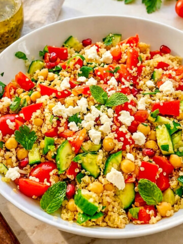 Une vue rapprochée et détaillée d'un bol de salade de quinoa et pois chiches mélangés, montrant les différentes textures des légumes coupés en dés, de la féta émiettée, du persil et des grains de grenade, avec une bouteille de vinaigrette à l'arrière-plan.