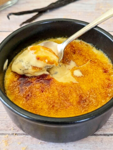 Crème brûlée traditionnelle à la vanille dans un ramequin noir avec une cuillère soulevant la croûte caramélisée.