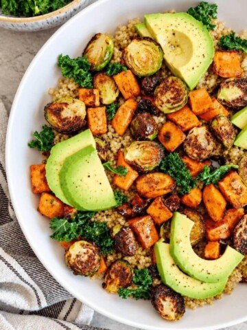 Salade de quinoa colorée dans un grand bol blanc, garnie de patates douces rôties, choux de Bruxelles grillés, chou kale et tranches d'avocat, servie sur un plan de travail en marbre clair.