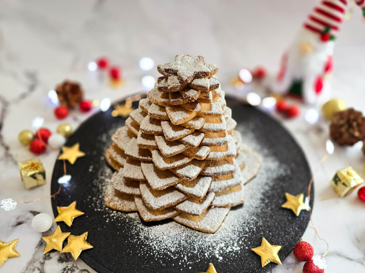 Un sapin de Noel en biscuits étoilés recouvert de sucre glace blanc, sur une assiette noire avec des décorations de Noel sur un fond blanc marbré.