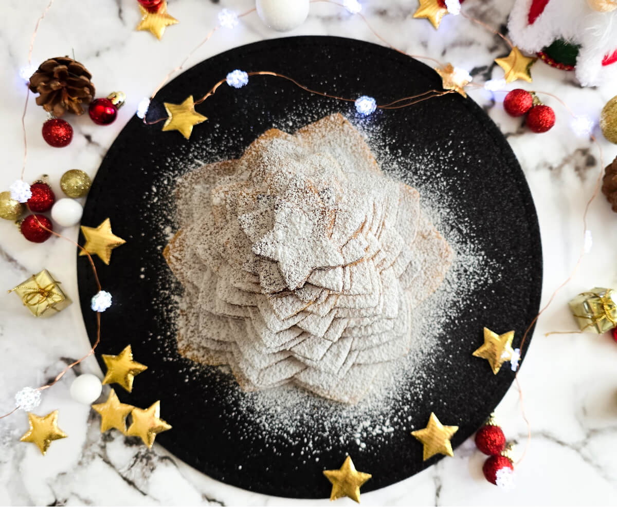 Un sapin de Noel en biscuits étoilés recouvert de sucre glace blanc, sur une assiette noire avec des décorations de Noel sur un fond blanc marbré.
