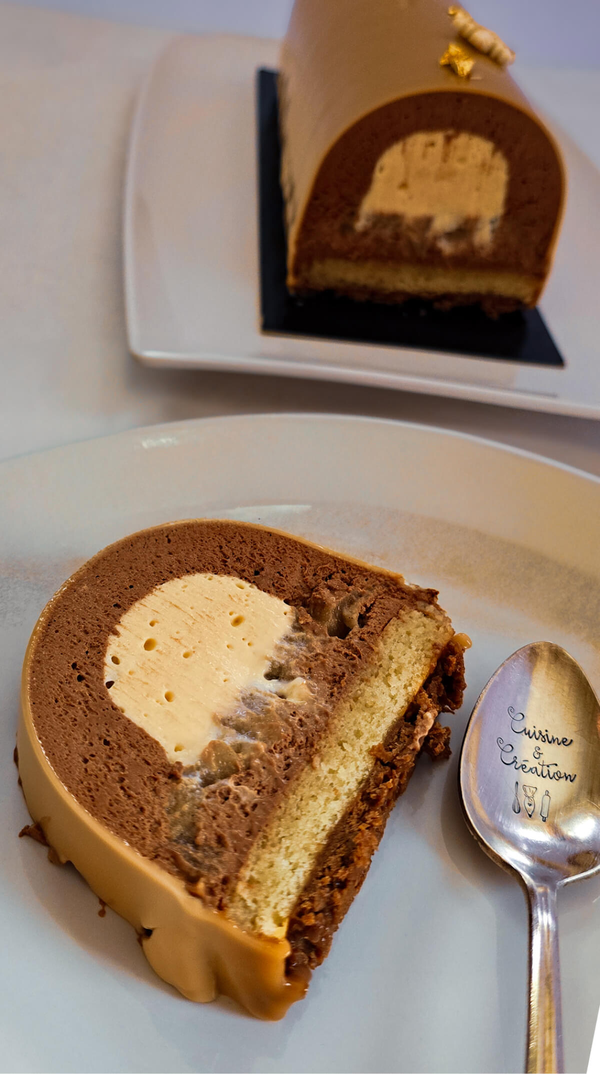 Une part de bûche de Noël poire caramel sur une assiette blanche avec une cuillère, avec la buche dans le fond.