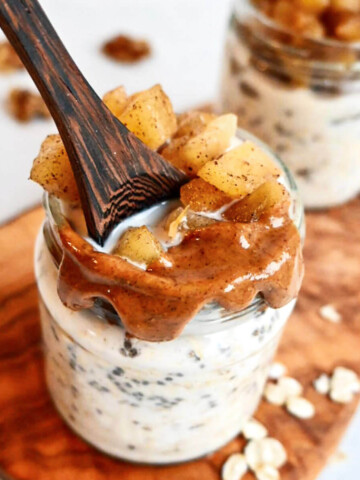 Deux contenants en verre avec une mixture blanche d'overnight oats, des pommes, une cuillère bois sur une planche de bois.