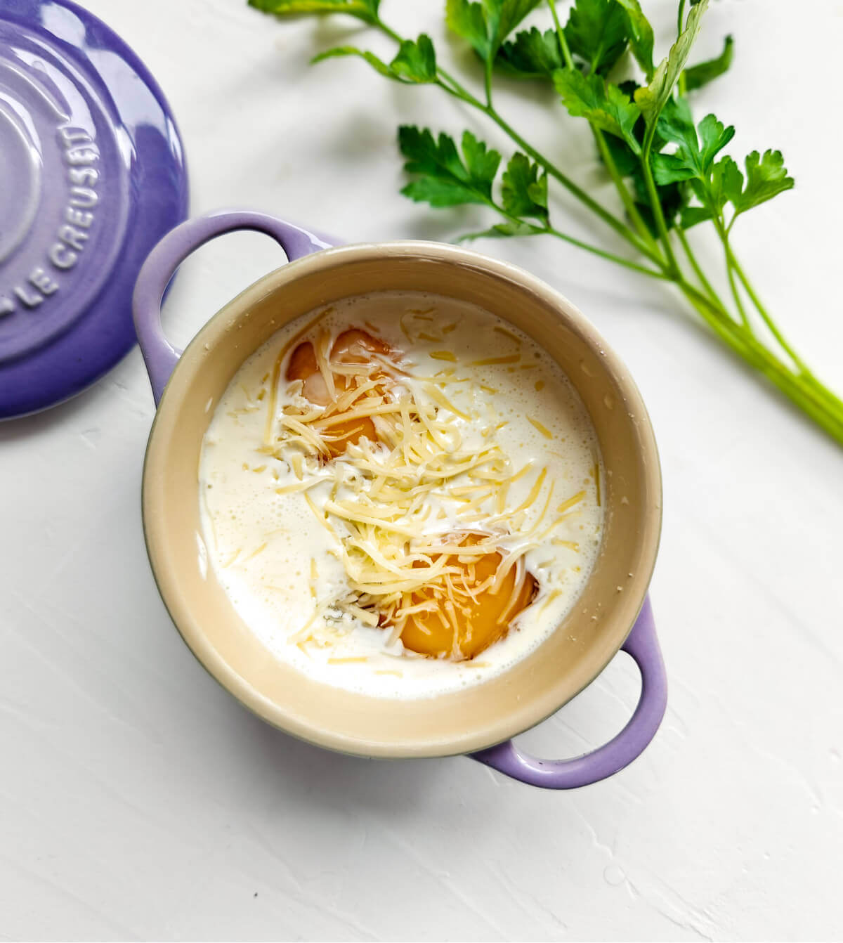 Deux oeufs non cuits dans une mini cocotte de couleur mauve sur un fond blanc. Persil sur le coté.