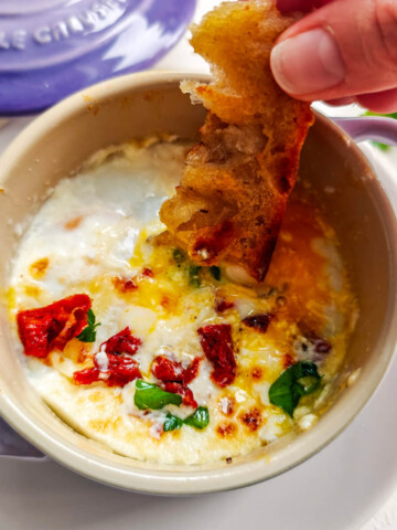 Des oeufs dans une cocotte indviduelle avec une main trempant un bout de pain.