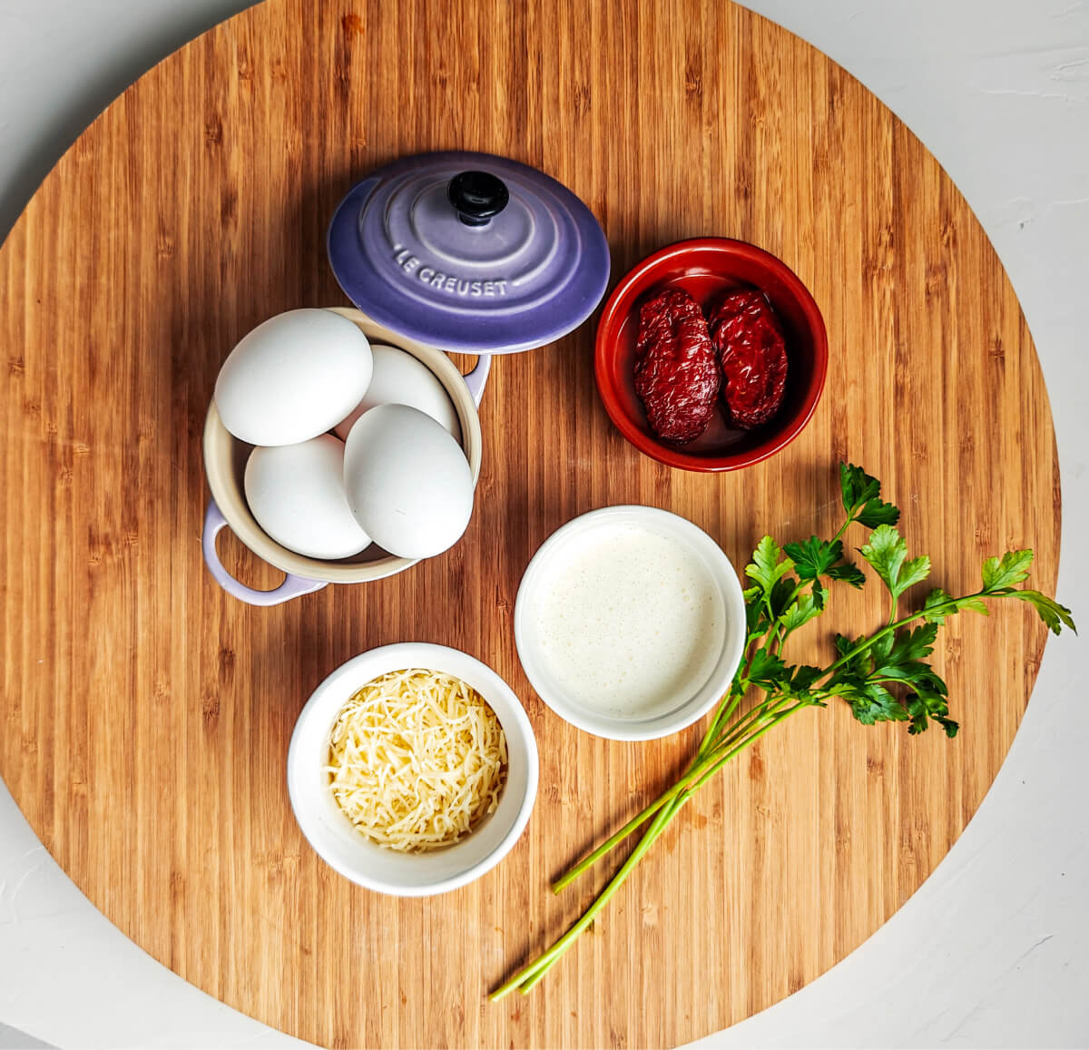 Des ingrédients pour une recette d'oeufs en cocotte, sur une planche de bois ronde, sur un fond blanc.