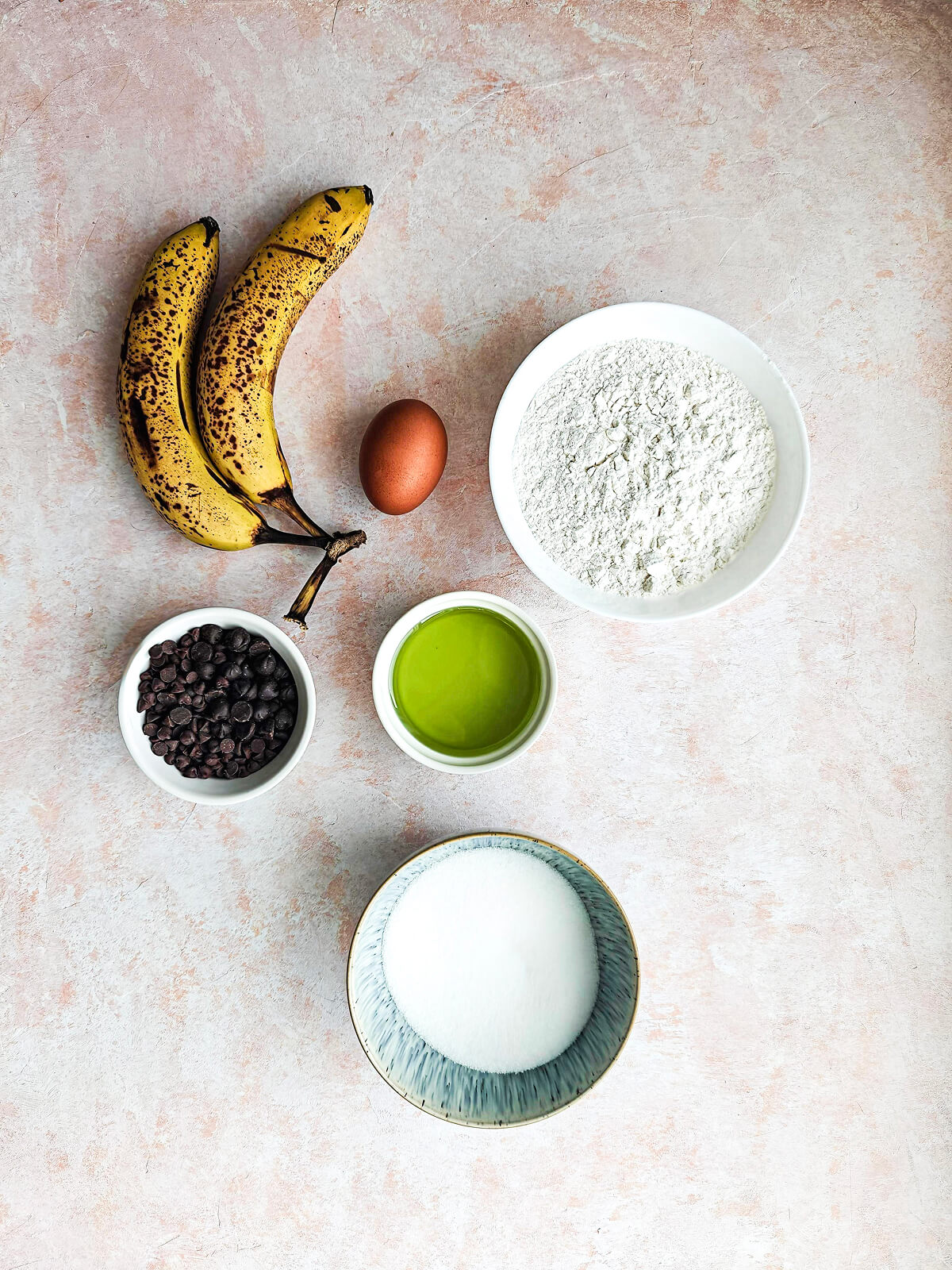 Des ingrédients pour une recette de muffins banane chocolat sur un fond rose clair.
