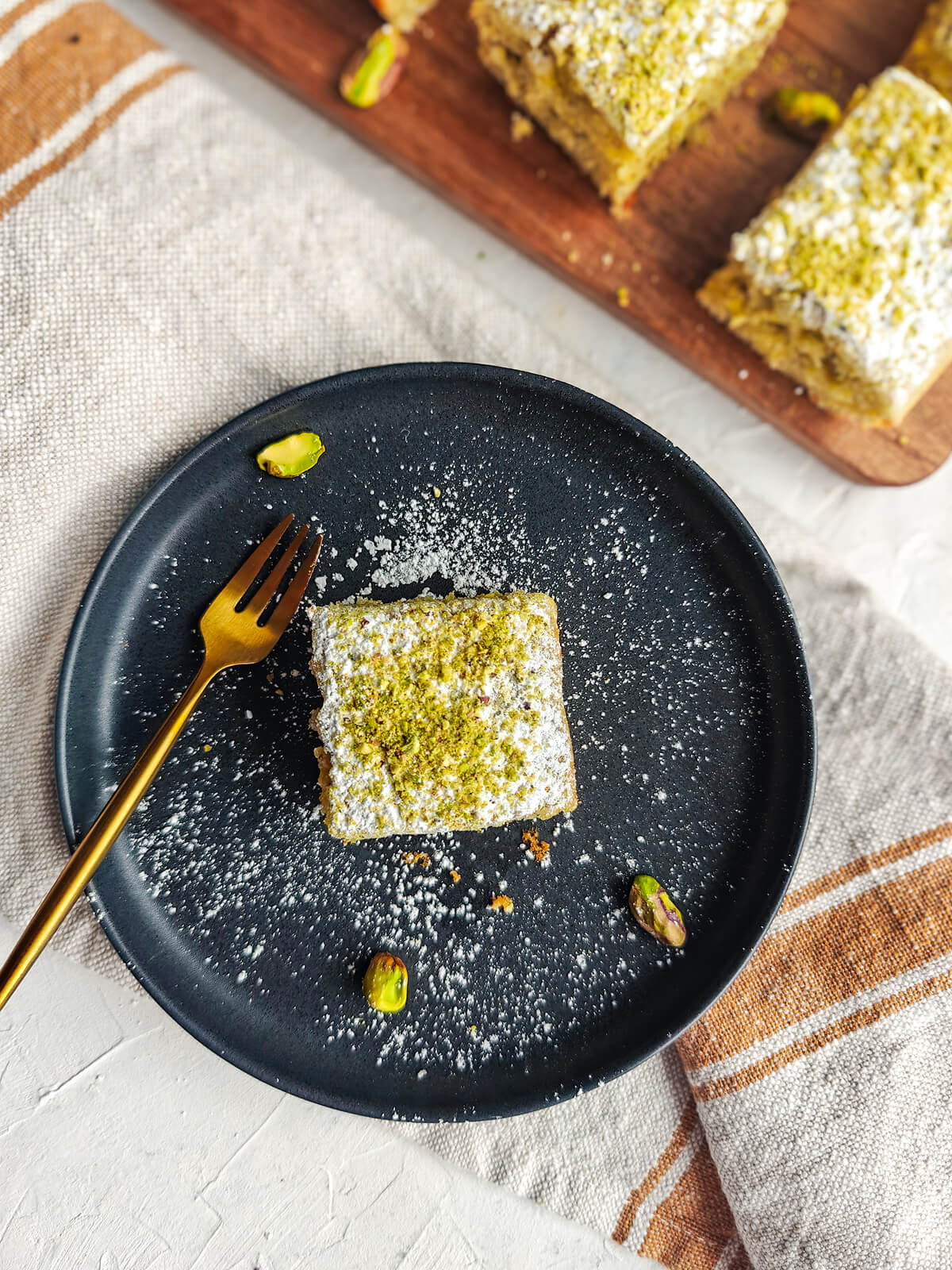 Une part de gateau dans une assiette noire avec une petite fourchette dorée, un torchon beige et quelques carrés de gateau sur le coté.