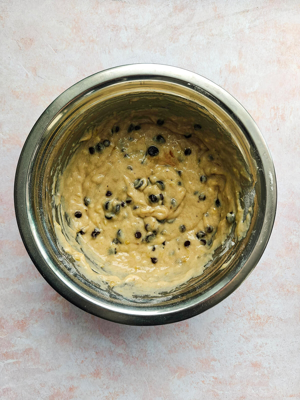 Une pâte à muffins avec pépites de chocolat dans un bol en acier inoxydable sur un fond rose clair.