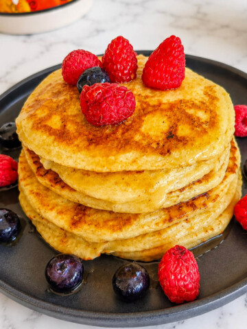 Des pancakes protéinés sans whey, bien fluffy, empilés les uns sur les autres, framboises sur le dessus et côté, sur une assiette noire, sur fond blanc marbré.