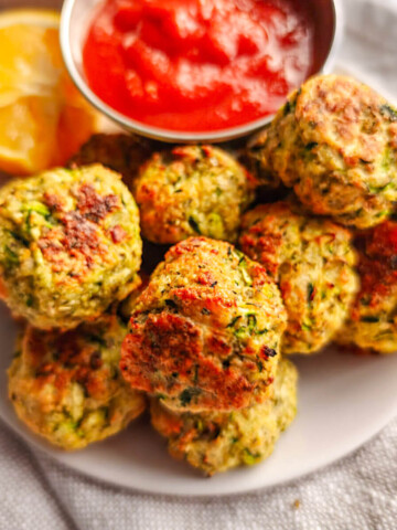 Des bouchées de courgettes apéro dans une assiette blanche avec un morceau de citron et de la sauce tomate dans un petit pot.