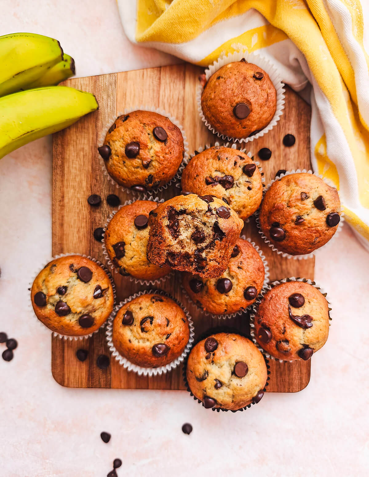 Des muffins sur une planche de bois, torchon jaune et bananes sur le coté, sur un fond rose clair.