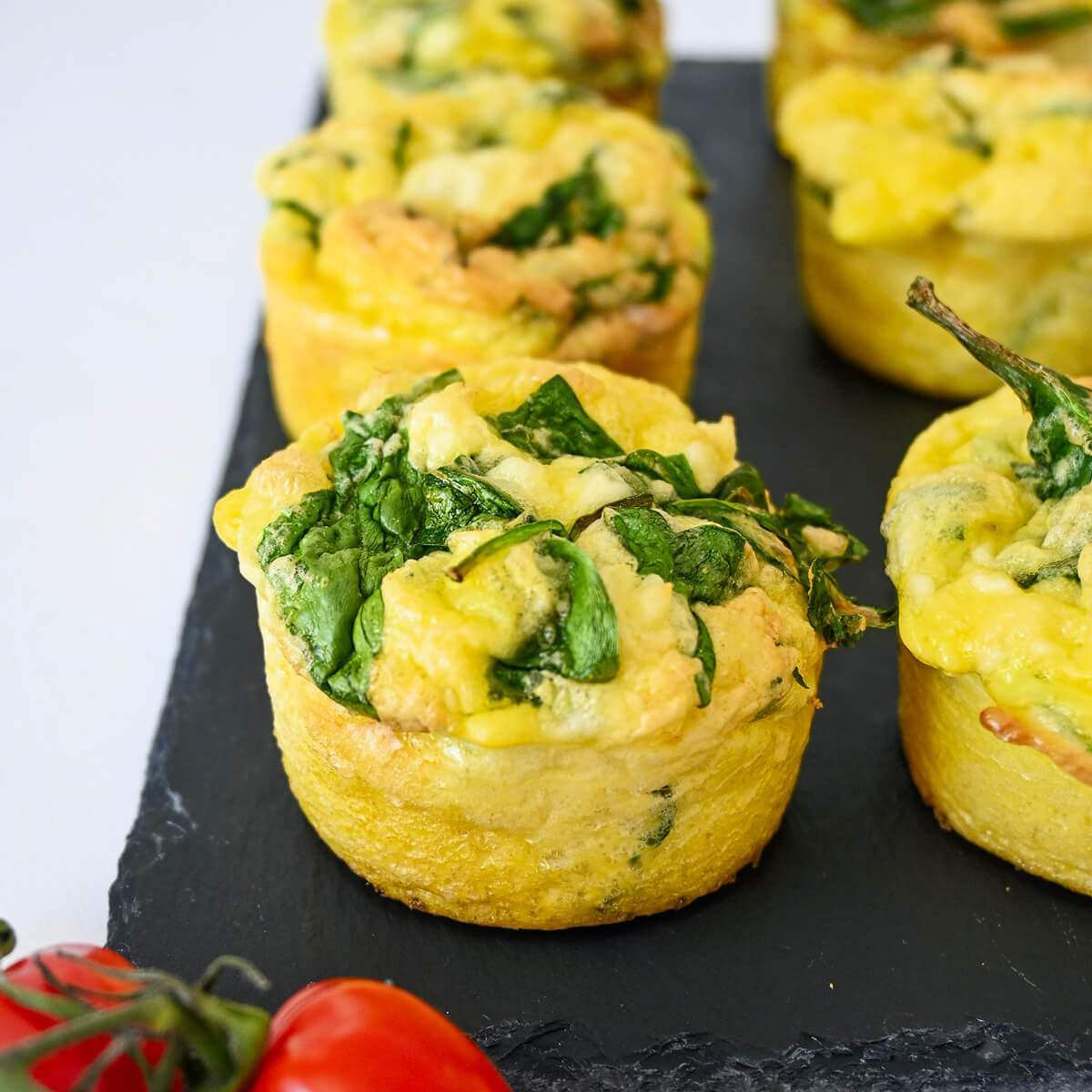 Des mini-quiches sans pâte épinards feta sur une assiette noire, tomates cerises sur le coté.