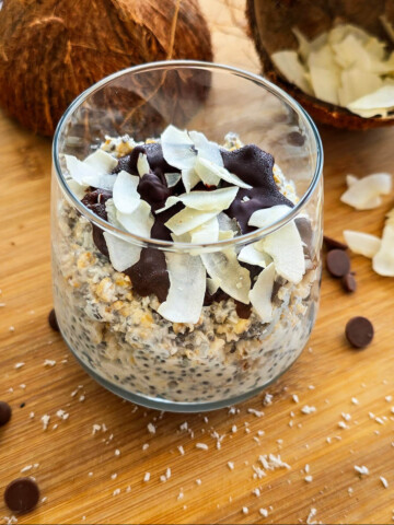 Un overnight porridge bounty dans un verre, sur une planche de bois, pépites de chocolat et copeaux de noix de coco sur le coté.