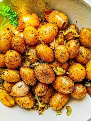Une assiette creuse de pommes de terre grenailles rôties à l'airfryer, bien dorées avec des herbes de Provence, du persil frais et des gousses d'ail en chemise.