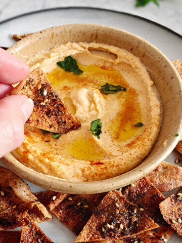 Un houmous bien crémeux dans un bol, avec une main tenant une chips de pita.