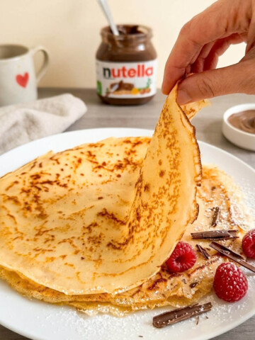 Une main tenant une crêpe sans gluten du bout des doigts, avec une assiette blanche, du Nutella et un café en arrière-plan.