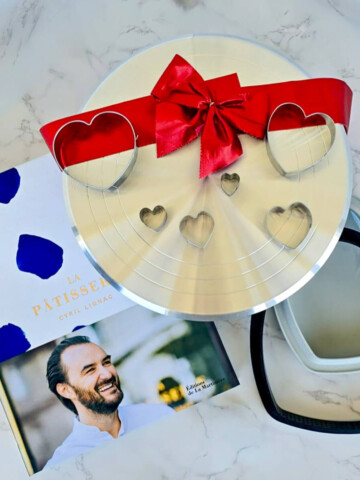 Sélection d'idées cadeaux pour la fête des mères : le livre de cuisine "La Pâtisserie" de Cyril Lignac, accompagné d'un moule à gâteau en forme de cœur et d'emporte-pièces assortis sur un plan de travail en marbre.