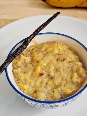 Bol de compote de banane à la vanille avec une gousse de vanille sur le rebord et une banane mûre en arrière-plan.