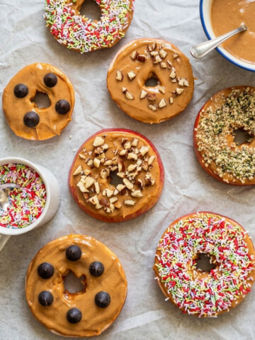 Vue de dessus de donuts de pommes (rondelles de pommes) nappées de beurre de cacahuète et décorées de pépites de chocolat, noisettes concassées, graines de chanvre et vermicelles colorés sur du papier cuisson.