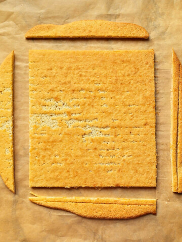 Un biscuit Joconde maison doré, découpé en un carré parfait sur une feuille de papier cuisson, avec les chutes de biscuit disposées sur les côtés.