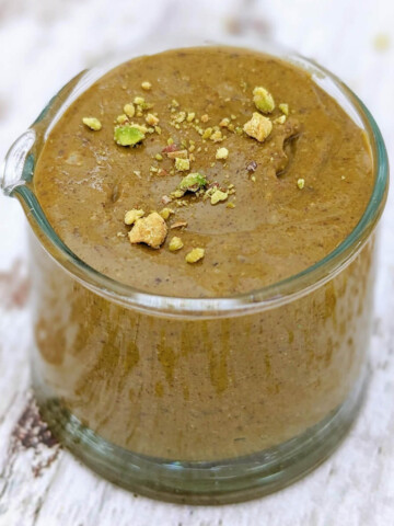 Du praliné pistache fait maison dans un petit contenant en verre, éclats de pistache concassées sur le dessus, sur un fond en bois clair.